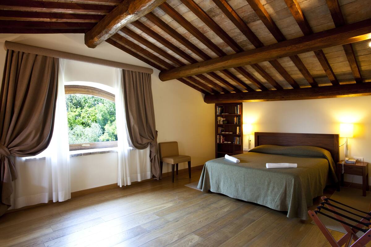 A bedroom with a wooden ceiling, a large window with curtains, a bed with a green bedspread, a bedside table with a lamp, and a bookshelf.