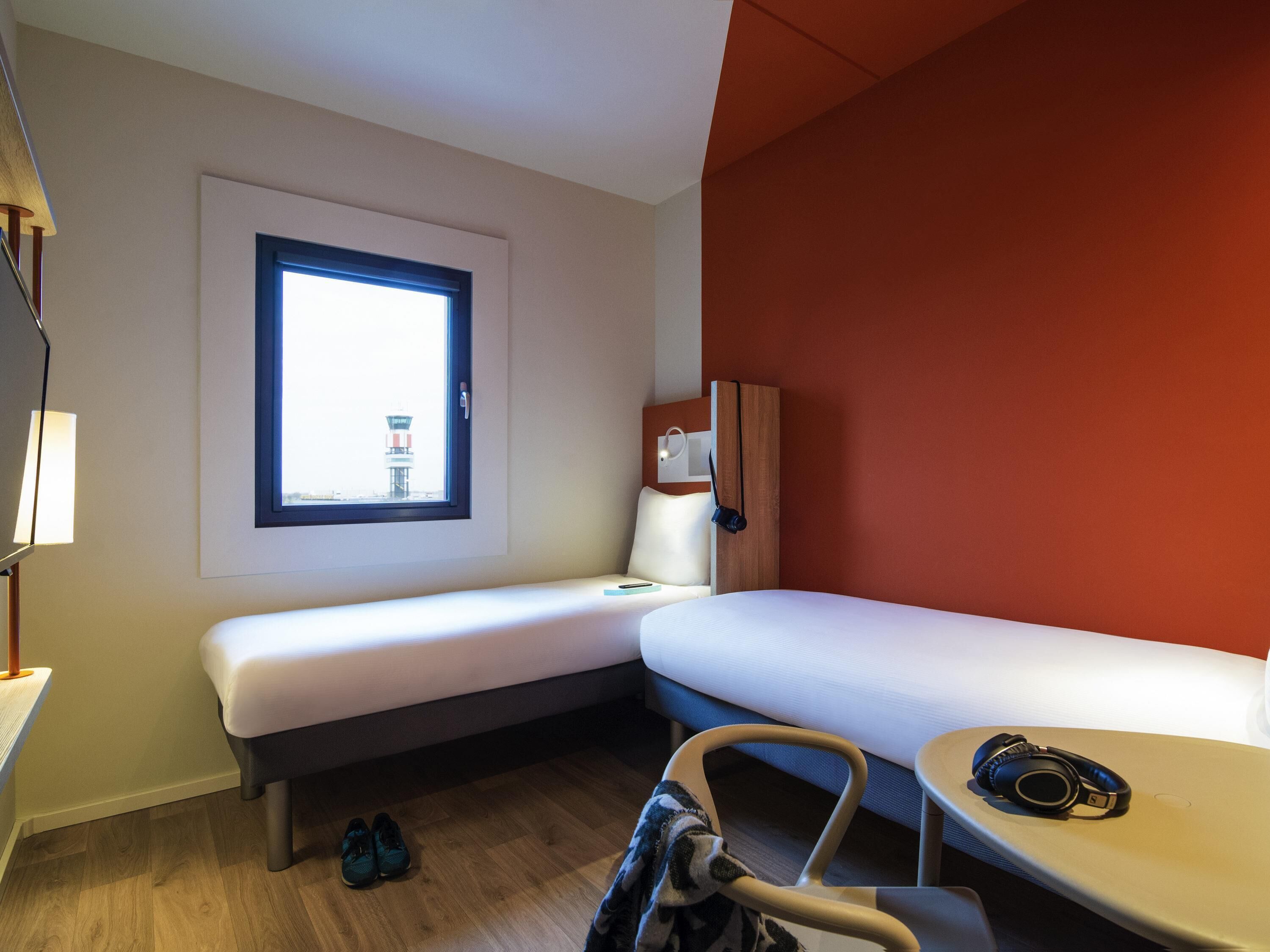 Une chambre d’hôtel avec un lit simple, un bureau, une chaise, une fenêtre donnant sur une tour et un mur d’accent rouge.