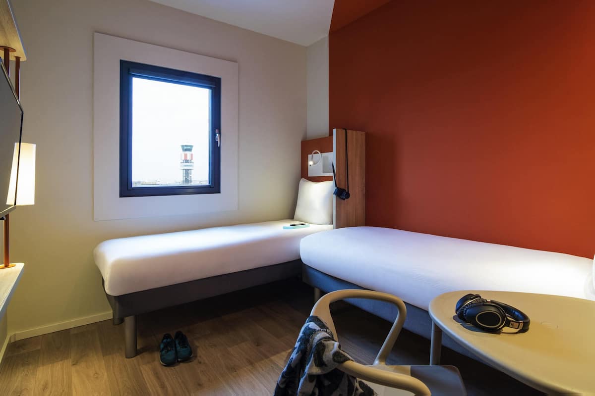 A hotel room with a single bed, a desk, a chair, a window with a view of a tower, and a red accent wall.