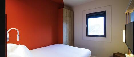 Ein Hotelzimmer mit Bett, Fenster, Fernseher und einem kleinen Hocker.
