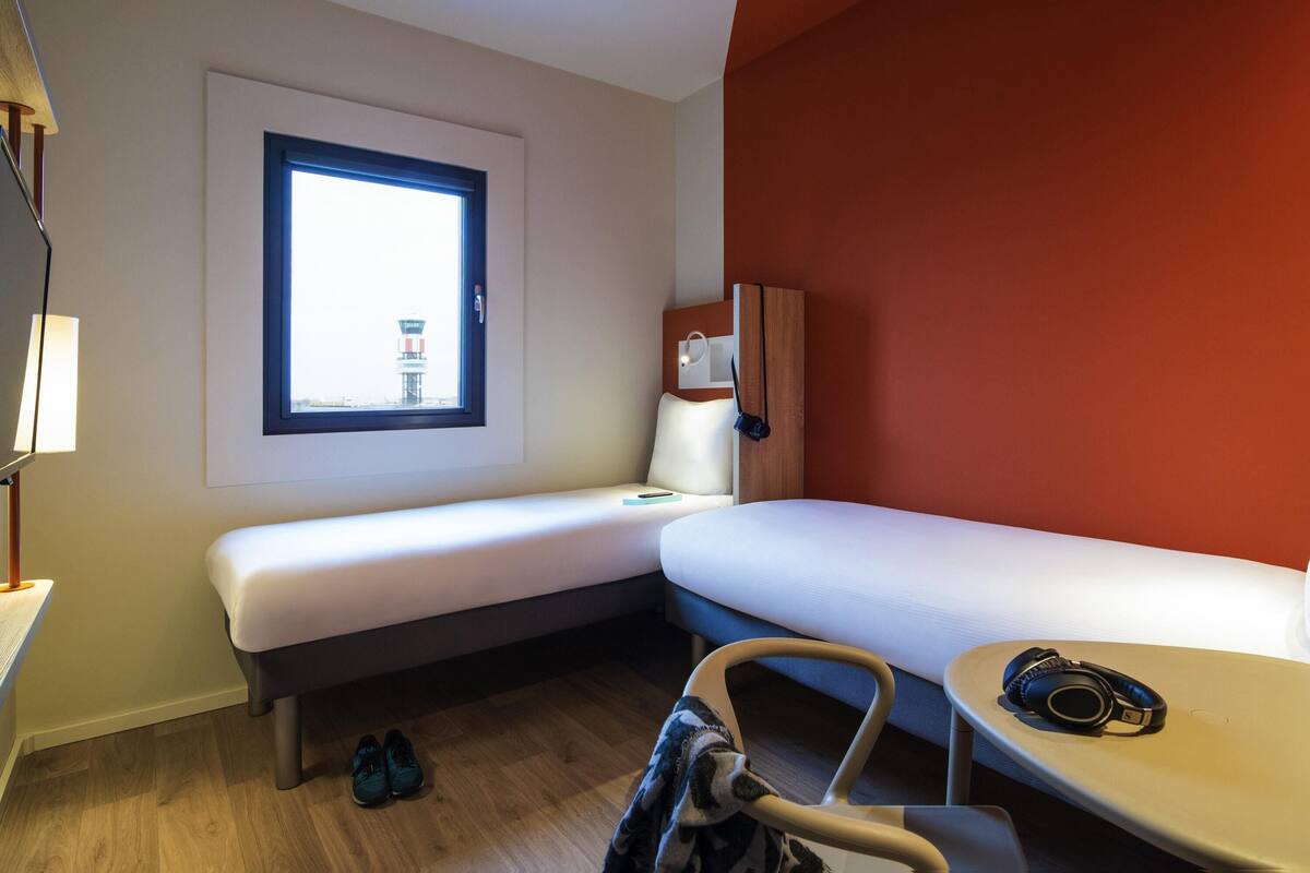 Habitación de hotel con una cama individual, un escritorio, una silla, un ventanal con vistas a una torre y una pared de acento roja.
