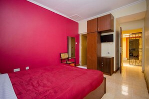 Une chambre avec un mur rouge, un lit, une armoire en bois, une télévision et une salle de bain visible à travers une porte ouverte.