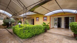 A covered courtyard with stone flooring, a green hedge, and a yellow building with red trim. - Konar House Kilimani (Nairobi)
