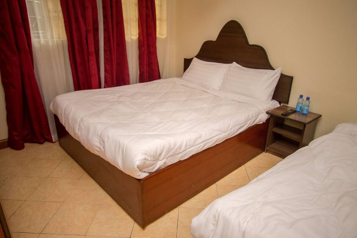 A hotel room with a large bed, a bedside table with two bottles, and red curtains.