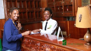 A hotel reception desk with a wooden finish, a clock, and a sign indicating the hotel is an embassy. - Hotel Embassy (Nairobi)