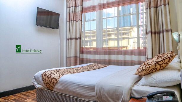 A hotel room with a bed, a TV mounted on the wall, a telephone, and a window with curtains. - Hotel Embassy (Nairobi)