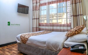 A hotel room with a bed, a TV mounted on the wall, a telephone, and a window with curtains. - Hotel Embassy (Nairobi)