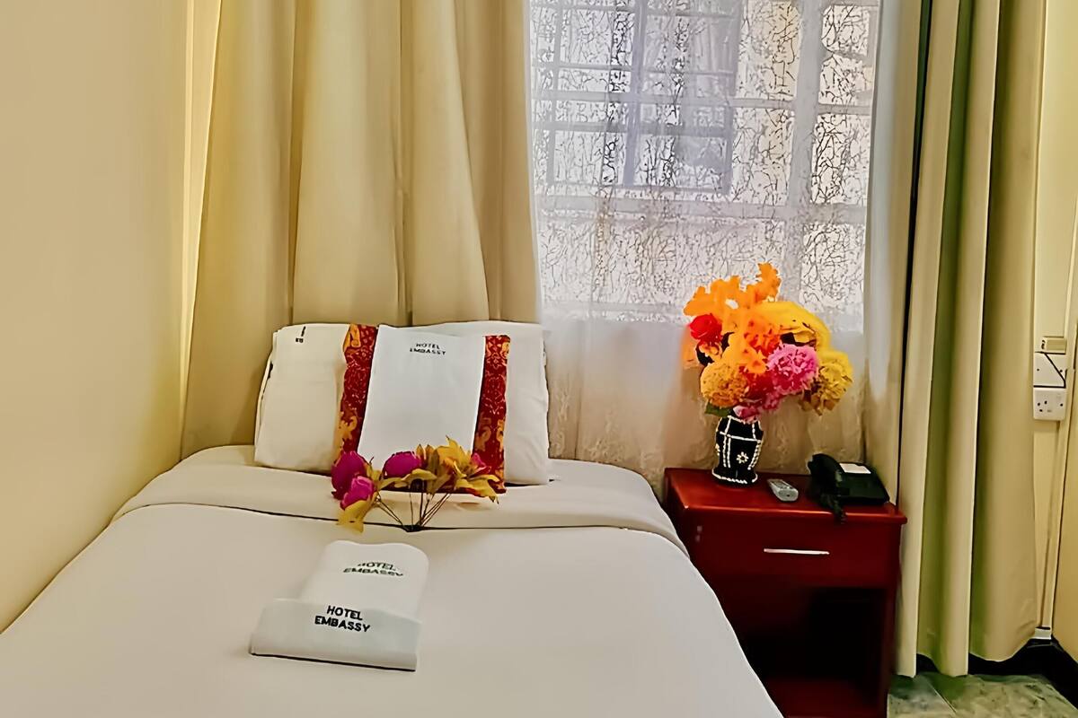 A hotel room with a bed, a nightstand with a vase of flowers, and a window with curtains.