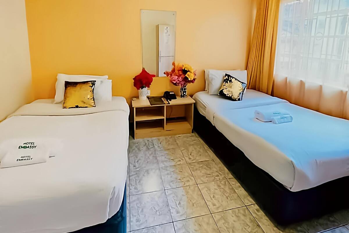 A hotel room with two beds, a small table with a vase of flowers, a mirror, and a window with curtains.