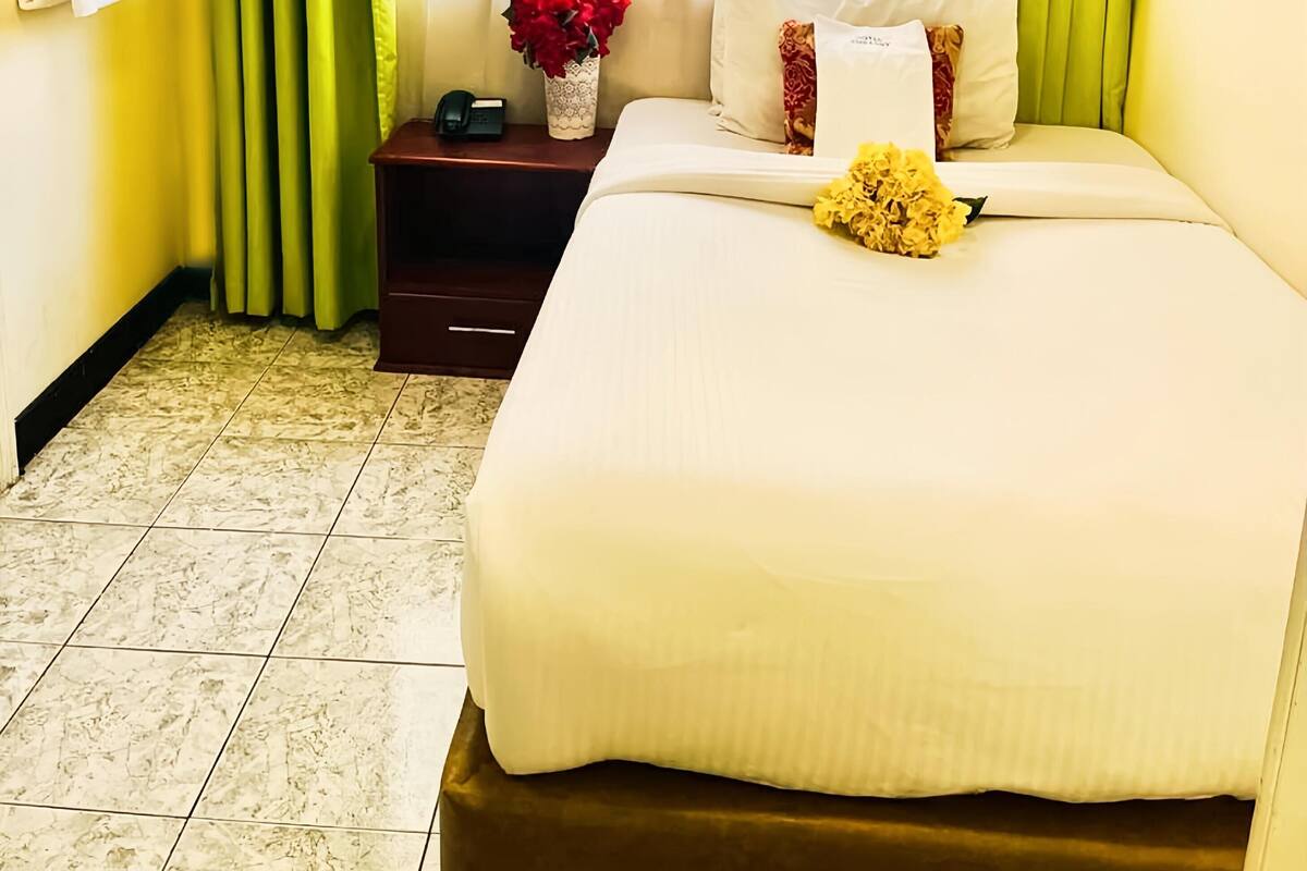 A hotel room with a bed, a nightstand, a vase with red flowers, and a window with green curtains.