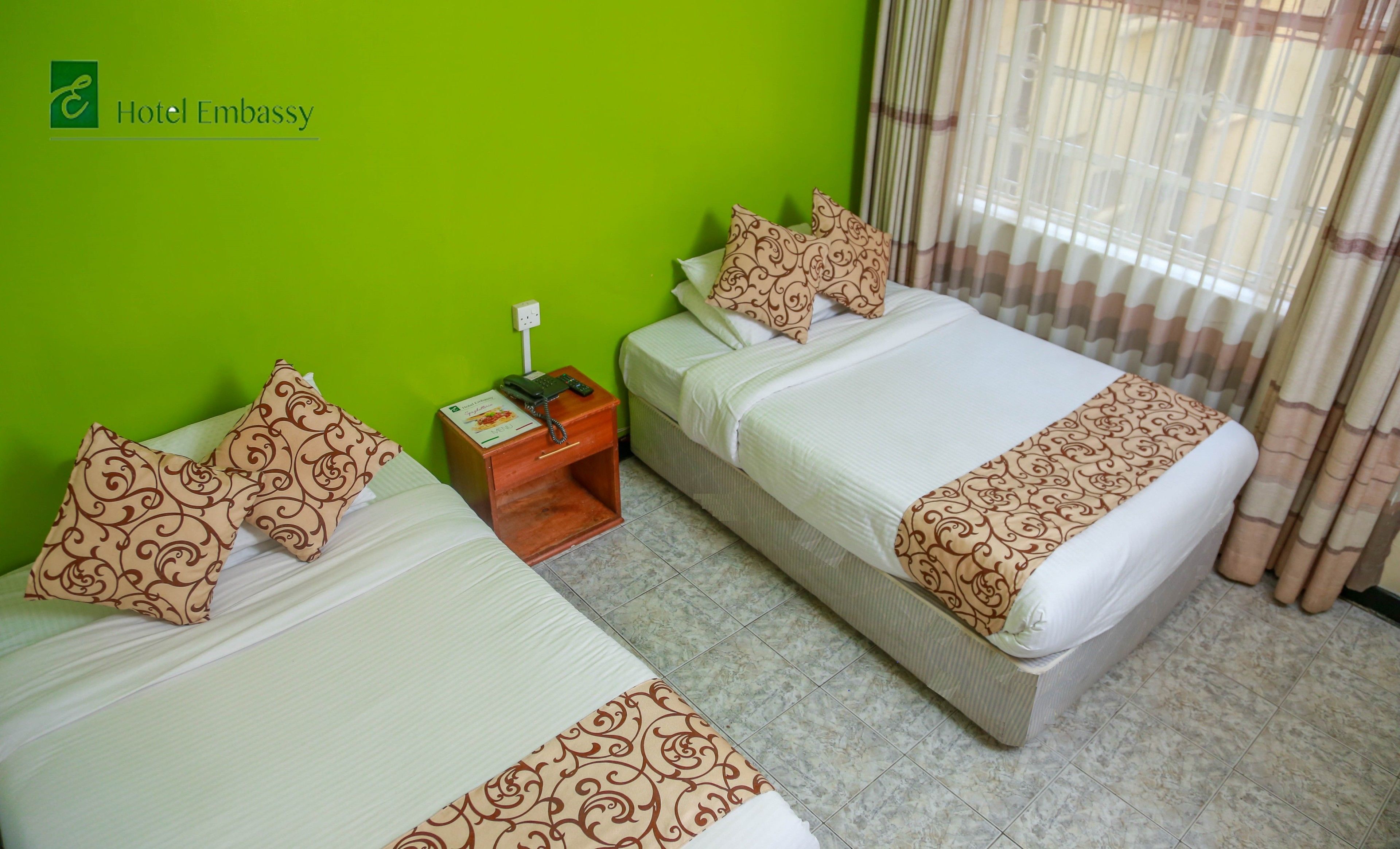 Habitación de hotel con dos camas, paredes verdes, una mesita de noche de madera con un teléfono y una ventana con cortinas.