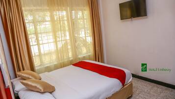 A hotel room with a bed, beige curtains, a TV on the wall, and a logo for Hotel Embassy.