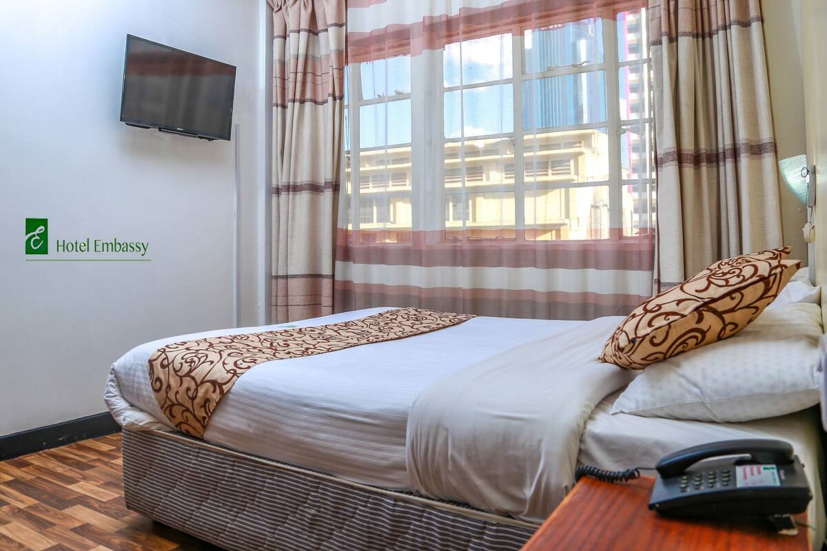 A hotel room with a bed, a TV mounted on the wall, a telephone, and a window with curtains.