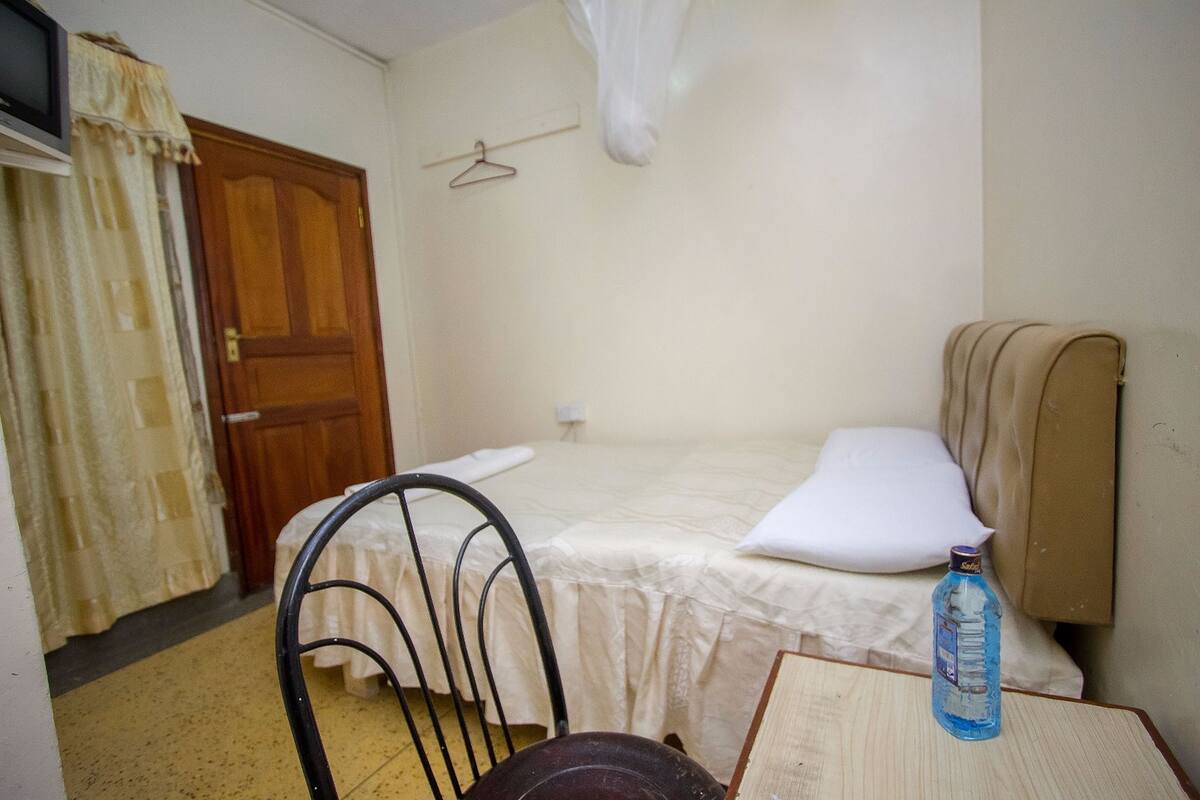 Quarto pequeno e simples com cama, cadeira, mesa e um cabide.