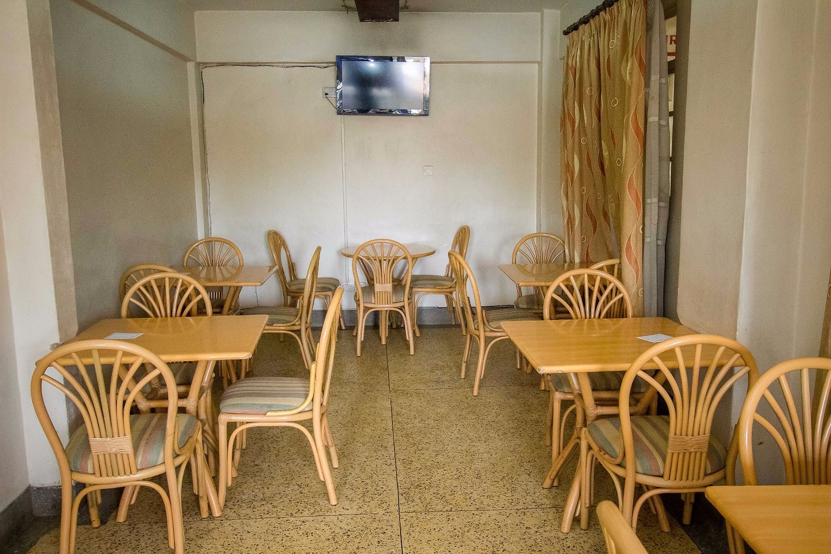 A room with wooden tables and chairs arranged in a U-shape, a television mounted on the wall, and a patterned curtain on the right side.
