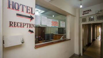 A hotel reception area with a reception desk, a window with various notices, and a hallway leading to other hotel rooms.