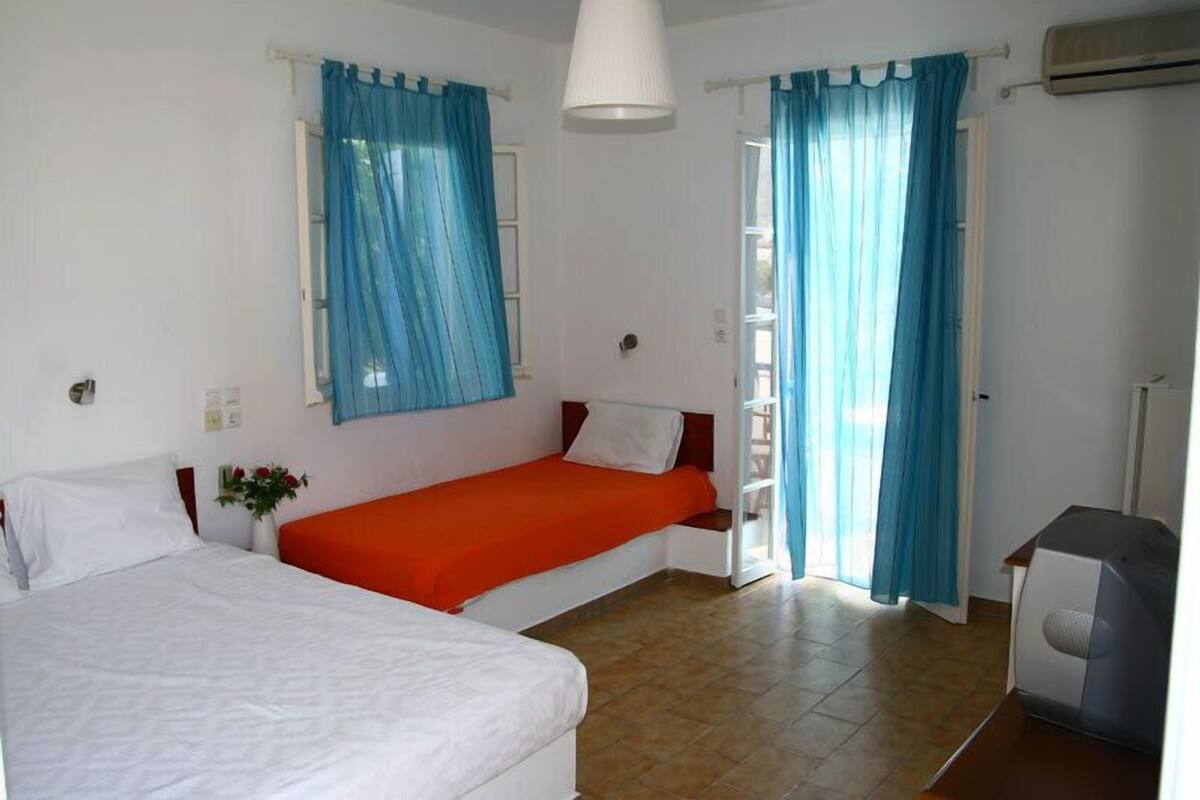 A room with two beds, one with an orange bedspread and the other with a white bedspread. There is a small table with a lamp, a window with blue curtains, and an air conditioning unit on the wall.