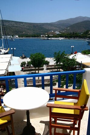 A balcony with a table and chairs overlooking a coastal area with boats and a mountainous backdrop. - Hotel Minoa (Amorgos)