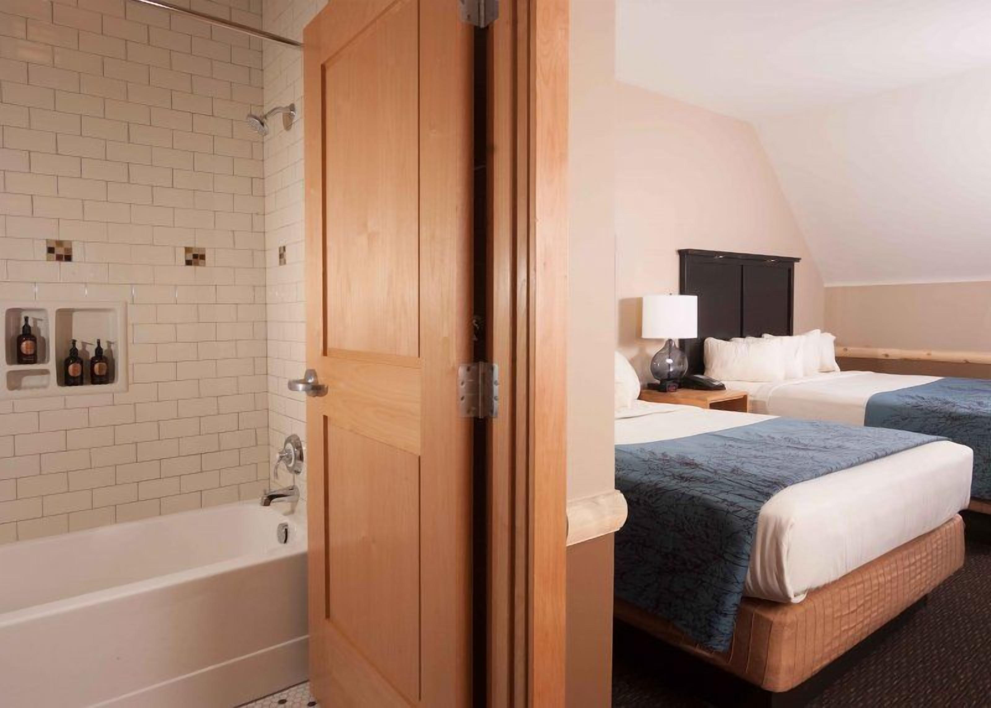 A hotel room with a wooden door, a bathtub, two beds with blue bedding, a nightstand with a lamp, and a wall-mounted shelf with bottles.