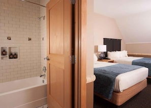 Quarto de hotel com porta de madeira, banheira, duas camas com roupa de cama azul, mesa de cabeceira com abajur e prateleira na parede com garrafas.