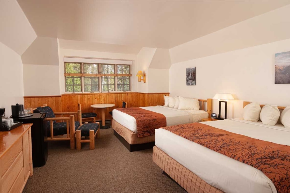 A hotel room with two beds, a small table, a chair, a desk, and a window with a view of trees.