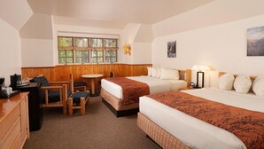 A hotel room with two beds, a small table, a chair, a desk, and a window with a view of trees.