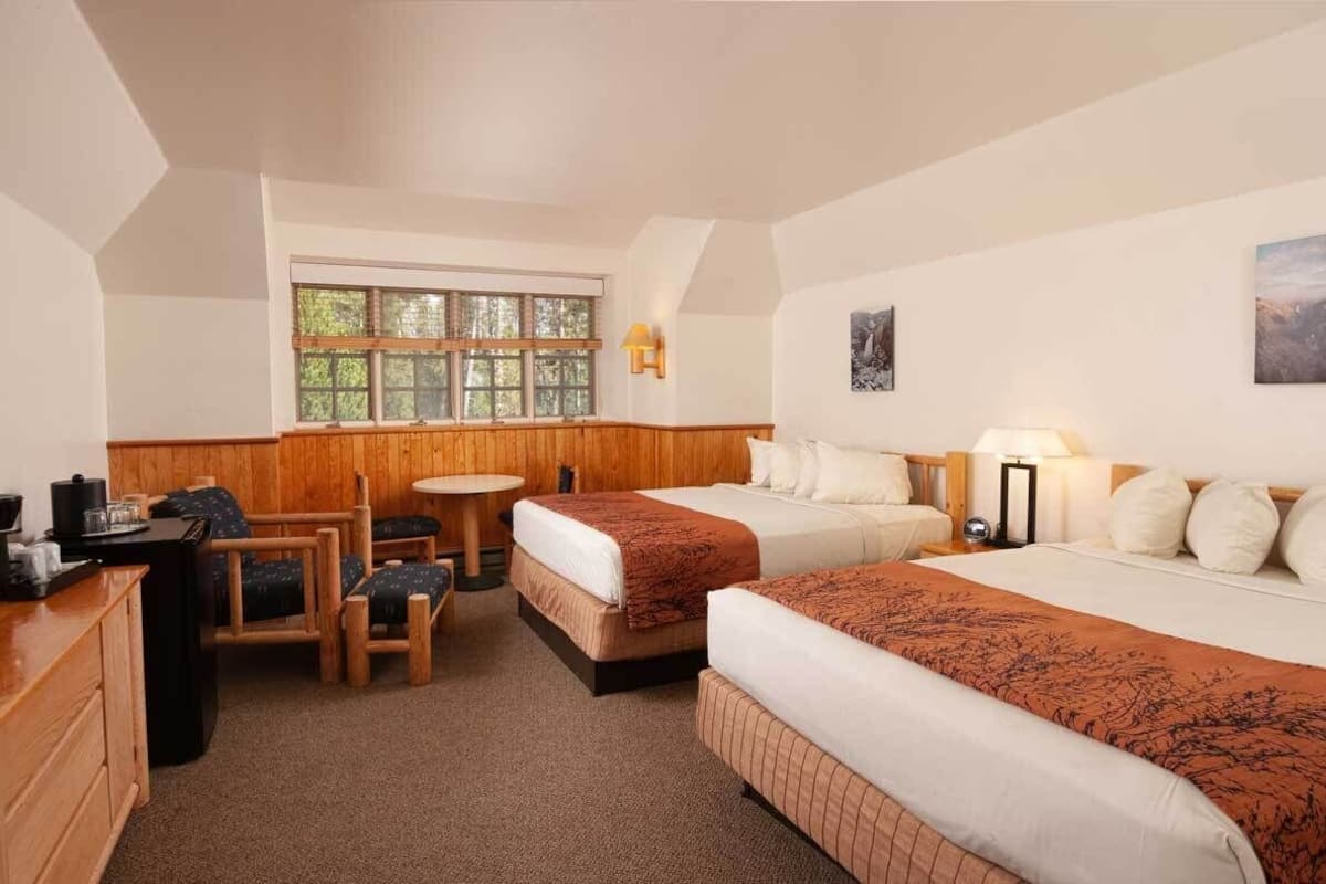 Ein Hotelzimmer mit zwei Betten, einem kleinen Tisch, einem Sessel, einem Schreibtisch und einem Fenster mit Blick auf Bäume.