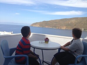 Two children seated at a small round table with a view of the sea and hills in the background. - Hotel Pelagos (Amorgos)
