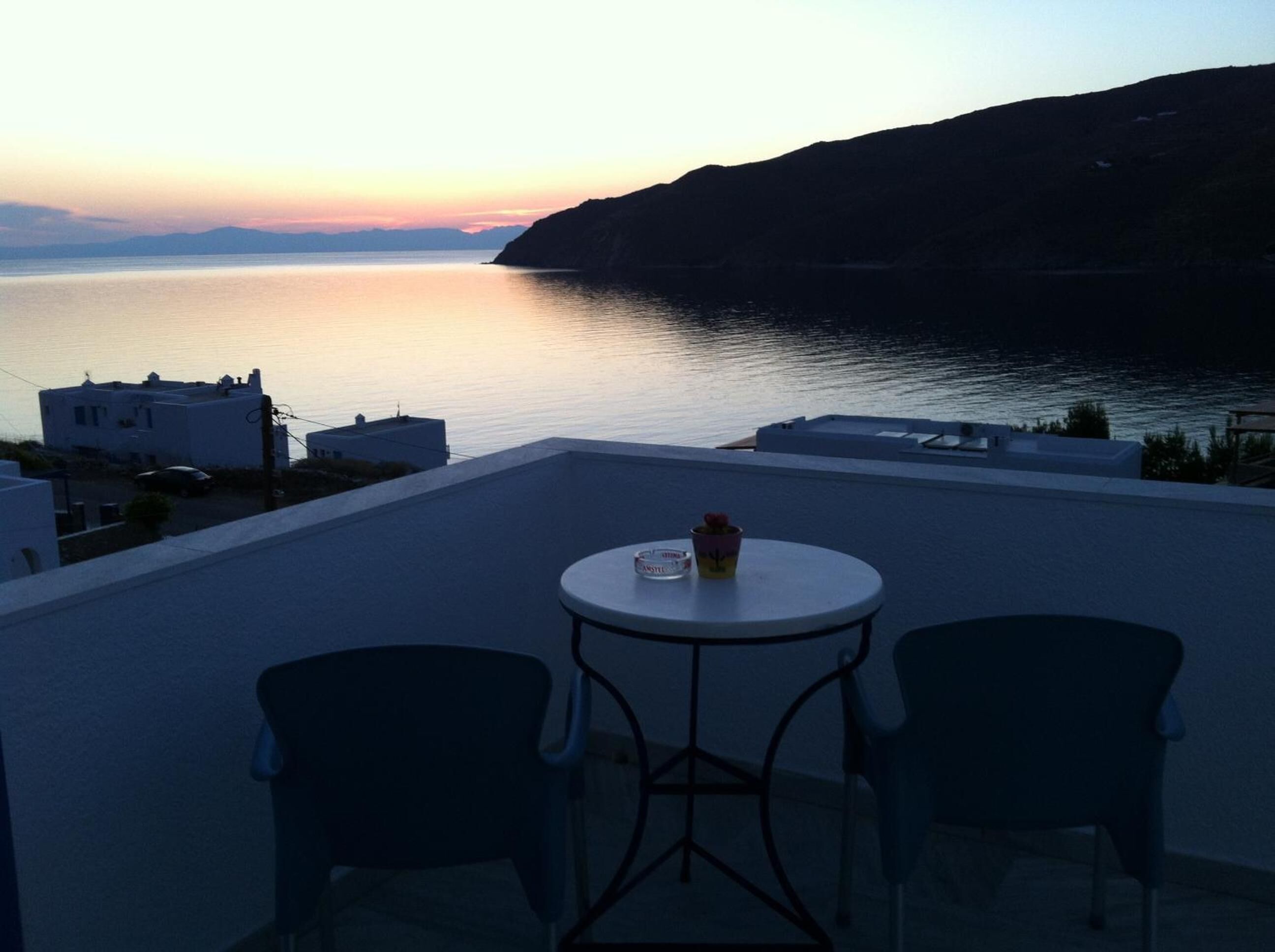 A terrace with two chairs and a table, overlooking a calm sea and a hilly landscape during sunset.