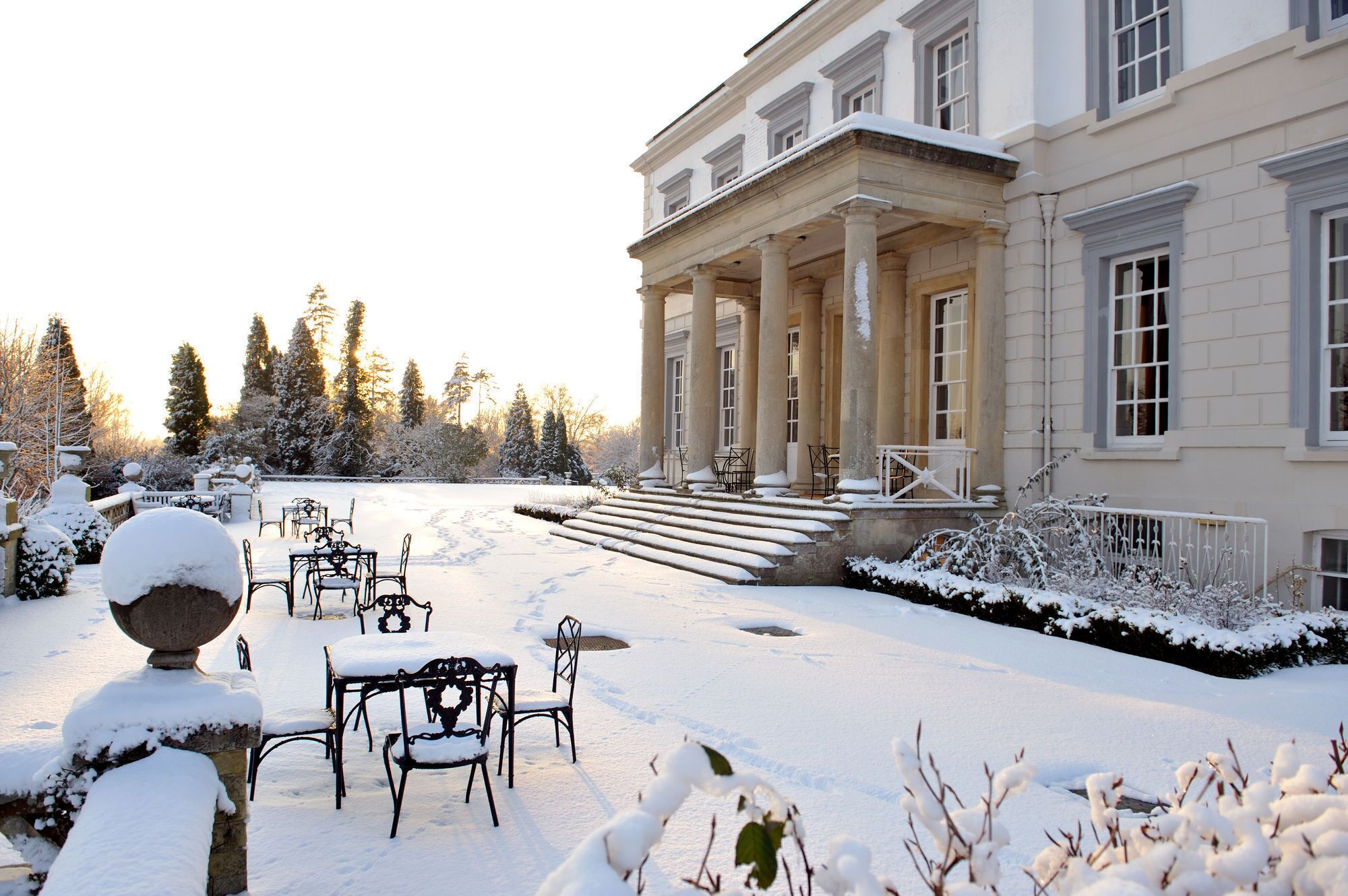 A snow-covered outdoor area with a classical building, steps leading to a porch, and a dining setup.