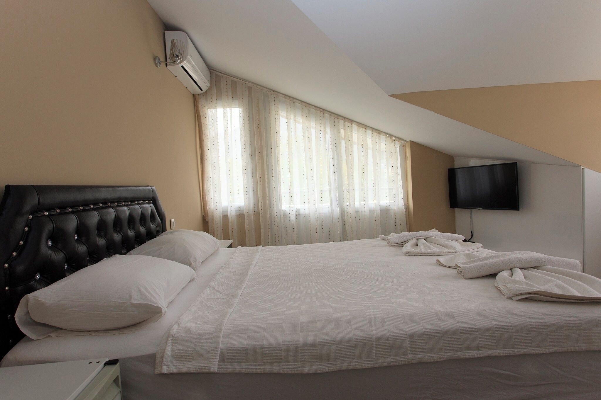 A hotel room with a large bed, a headboard, a flat-screen TV, and a window with curtains.