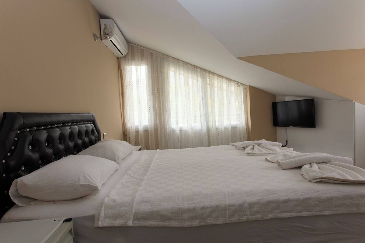 A hotel room with a large bed, a headboard, a flat-screen TV, and a window with curtains.