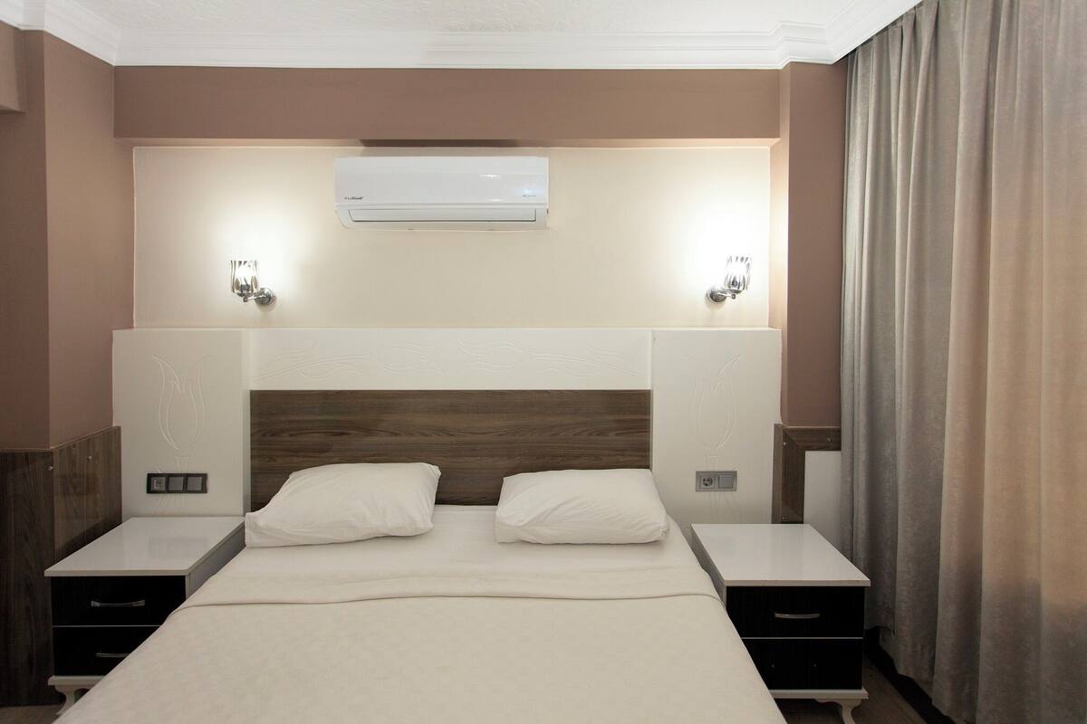 A hotel room with a large bed, two bedside tables, a wall-mounted air conditioner, and two wall sconces.