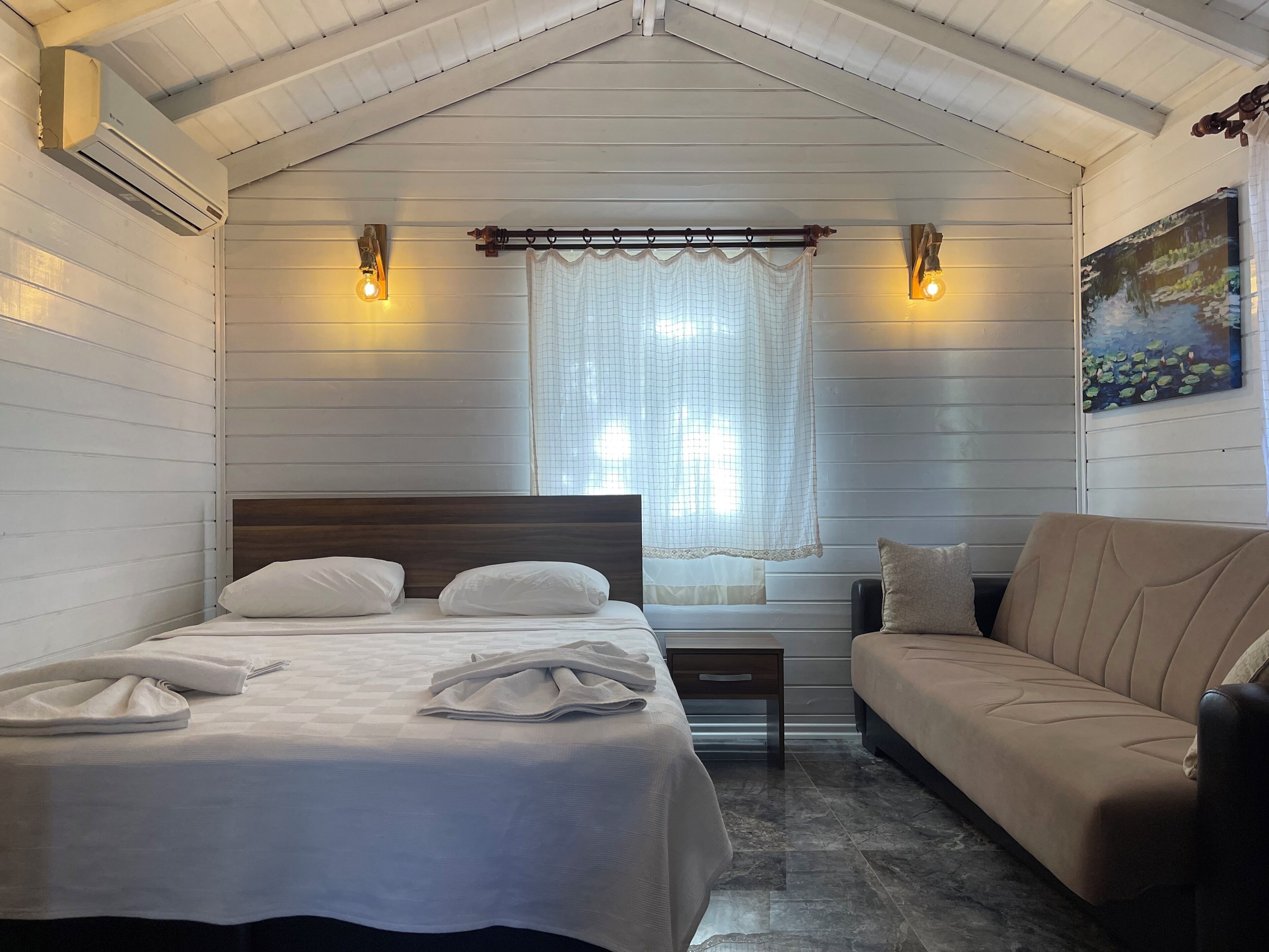 A bedroom with a bed, a sofa, a window with curtains, and wall-mounted lights.