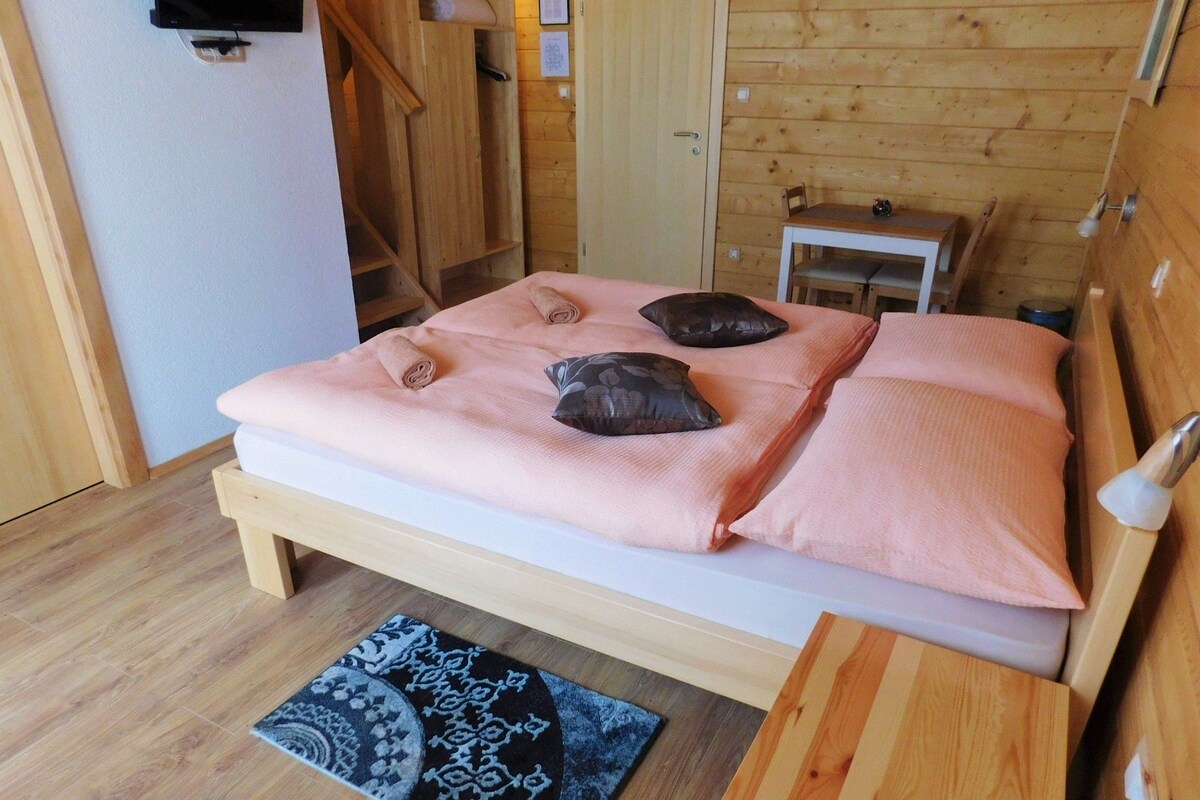 A wooden bed with a pink blanket and pillows, a small wooden table, a patterned rug, and a wooden wall.