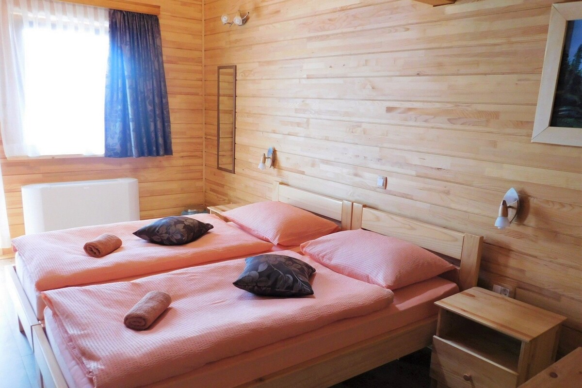A wooden cabin-style room with two beds, a small bedside table, a mirror, and a window with curtains.