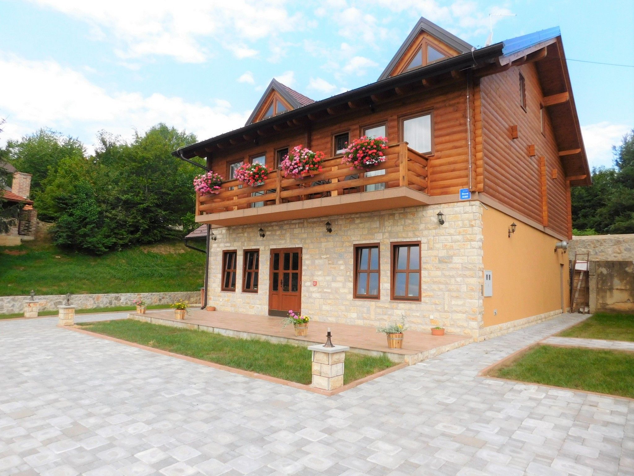 A two-story log cabin with a stone facade, wooden balcony with flower boxes, and a paved entrance area.