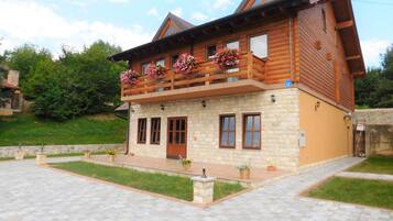 A two-story log cabin with a stone facade, wooden balcony with flower boxes, and a paved entrance area.