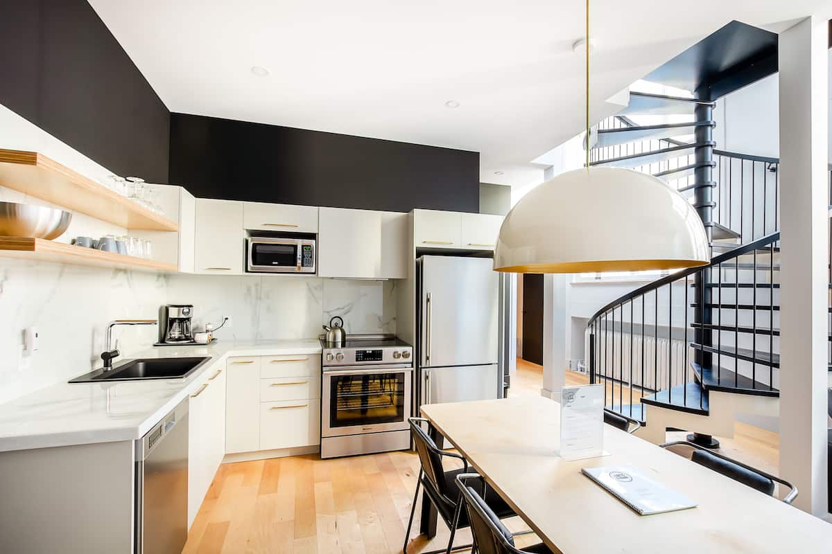 A modern kitchen with white cabinetry, a built-in oven, a microwave, and a stainless steel sink. There is a dining area with a table and chairs, and a black spiral staircase in the background.
