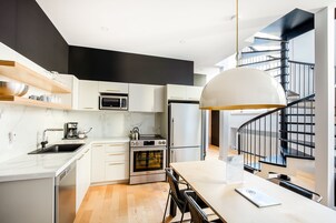 Una cocina moderna con armarios blancos, horno empotrado, microondas y pileta de acero inoxidable. Hay un comedor con mesa y sillas, y una escalera en espiral negra al fondo.