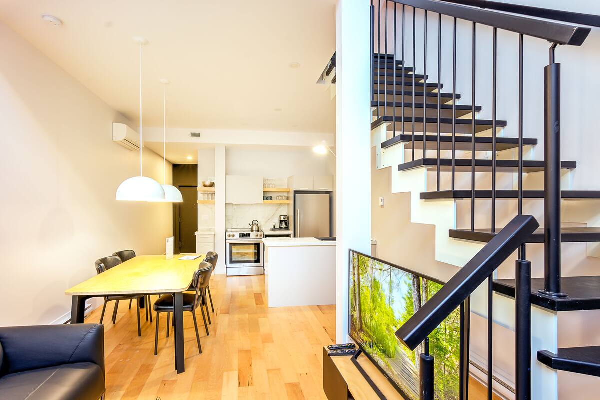 A modern kitchen with a central island, a dining area with a wooden table and chairs, and a staircase with black railings.