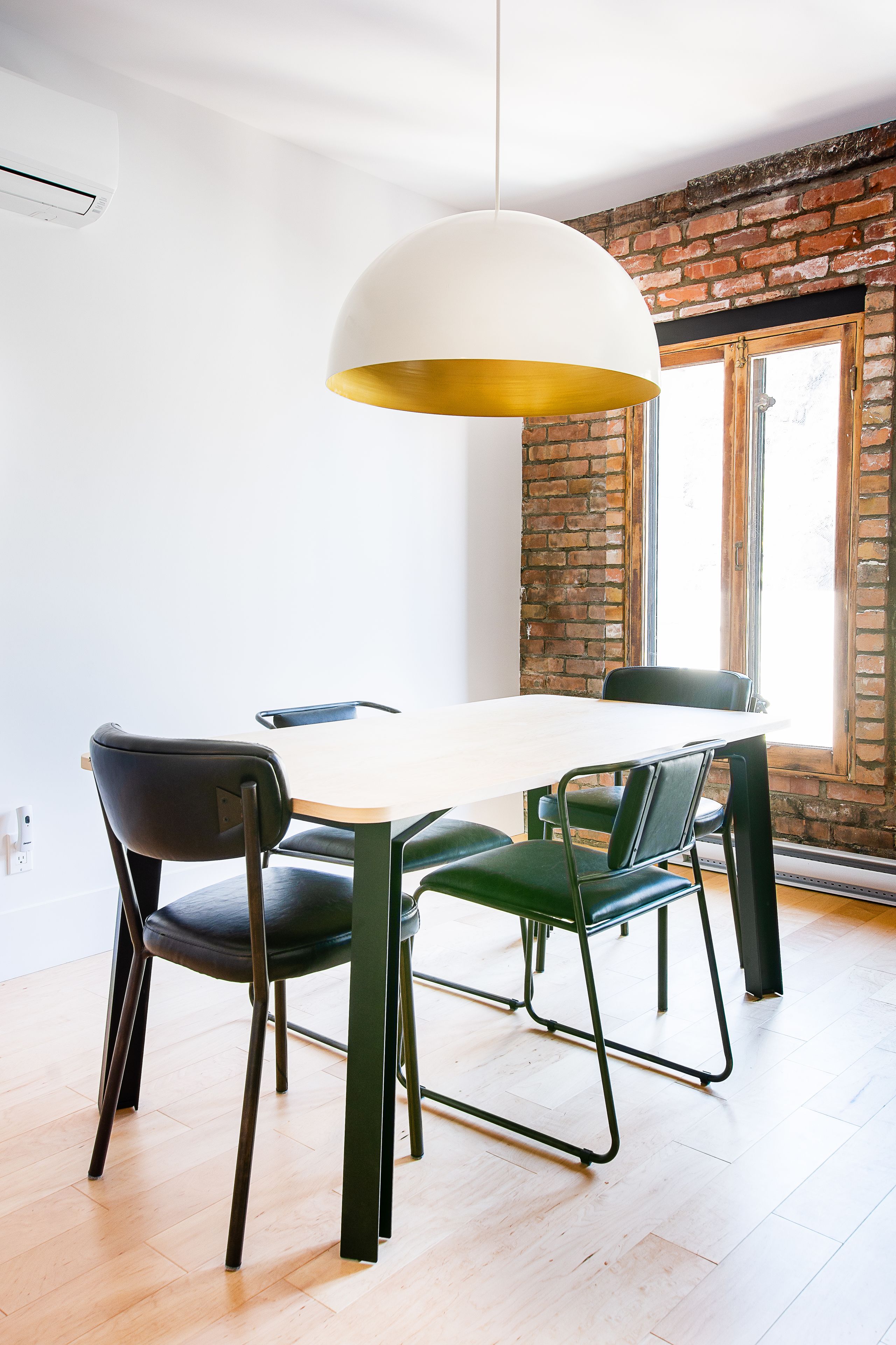 A dining area with a white table, black and green chairs, a white pendant light, and a brick wall with a window.