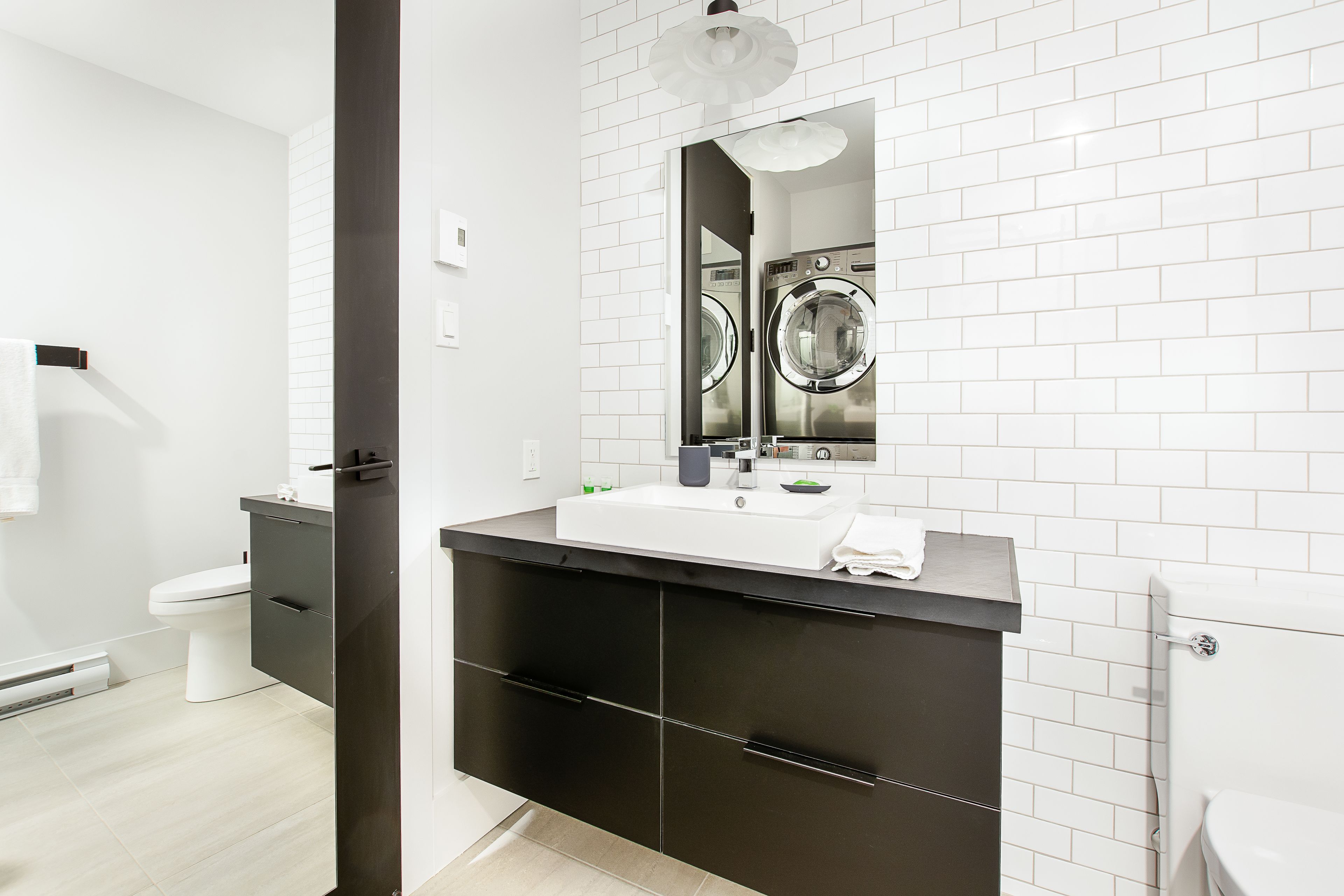 Un baño moderno con pared de azulejos blancos, un tocador negro con lavabo y dos lavadoras apiladas al fondo.