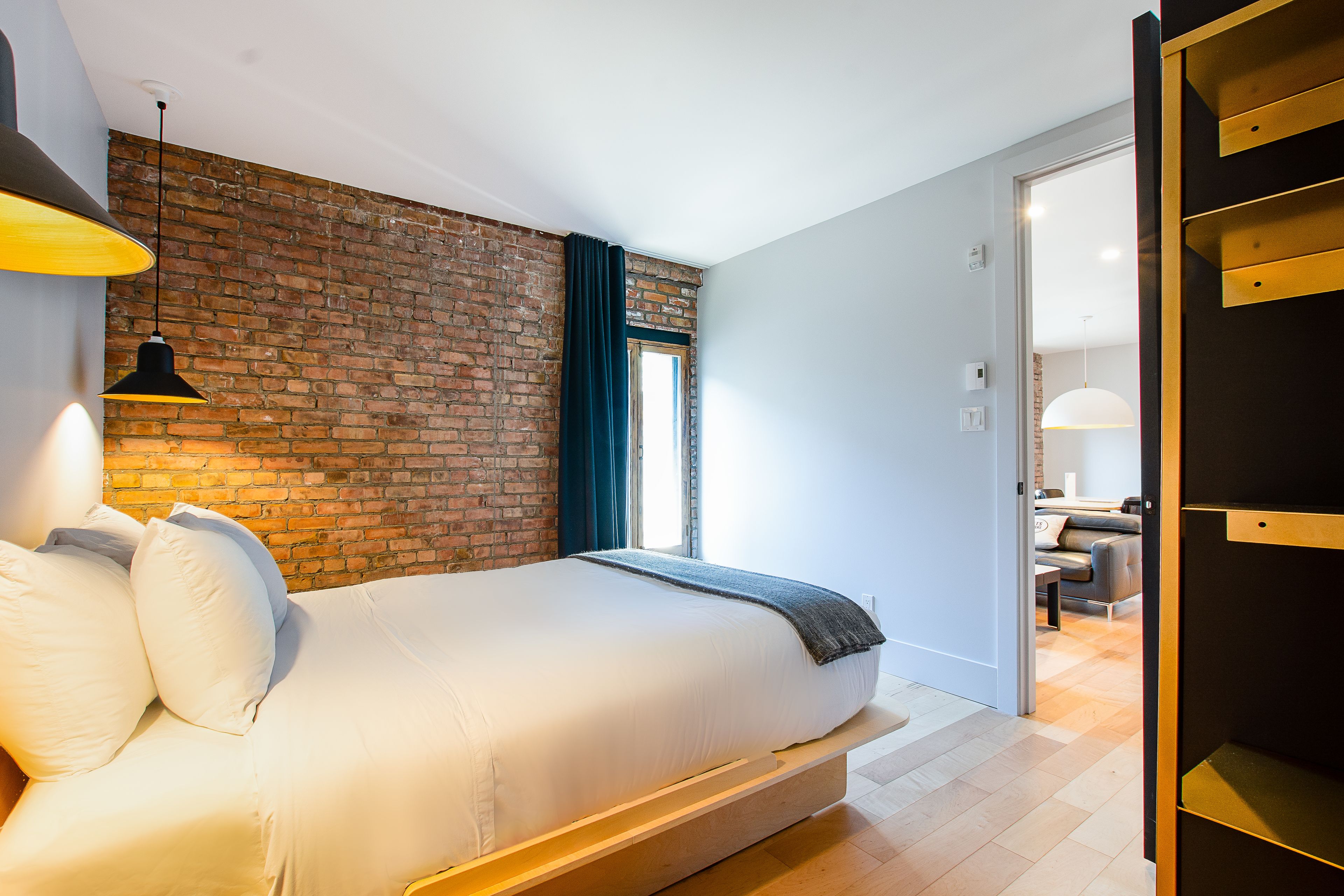 A modern bedroom with a brick wall, a large bed with white bedding, a hanging lamp, and a view into the living area.