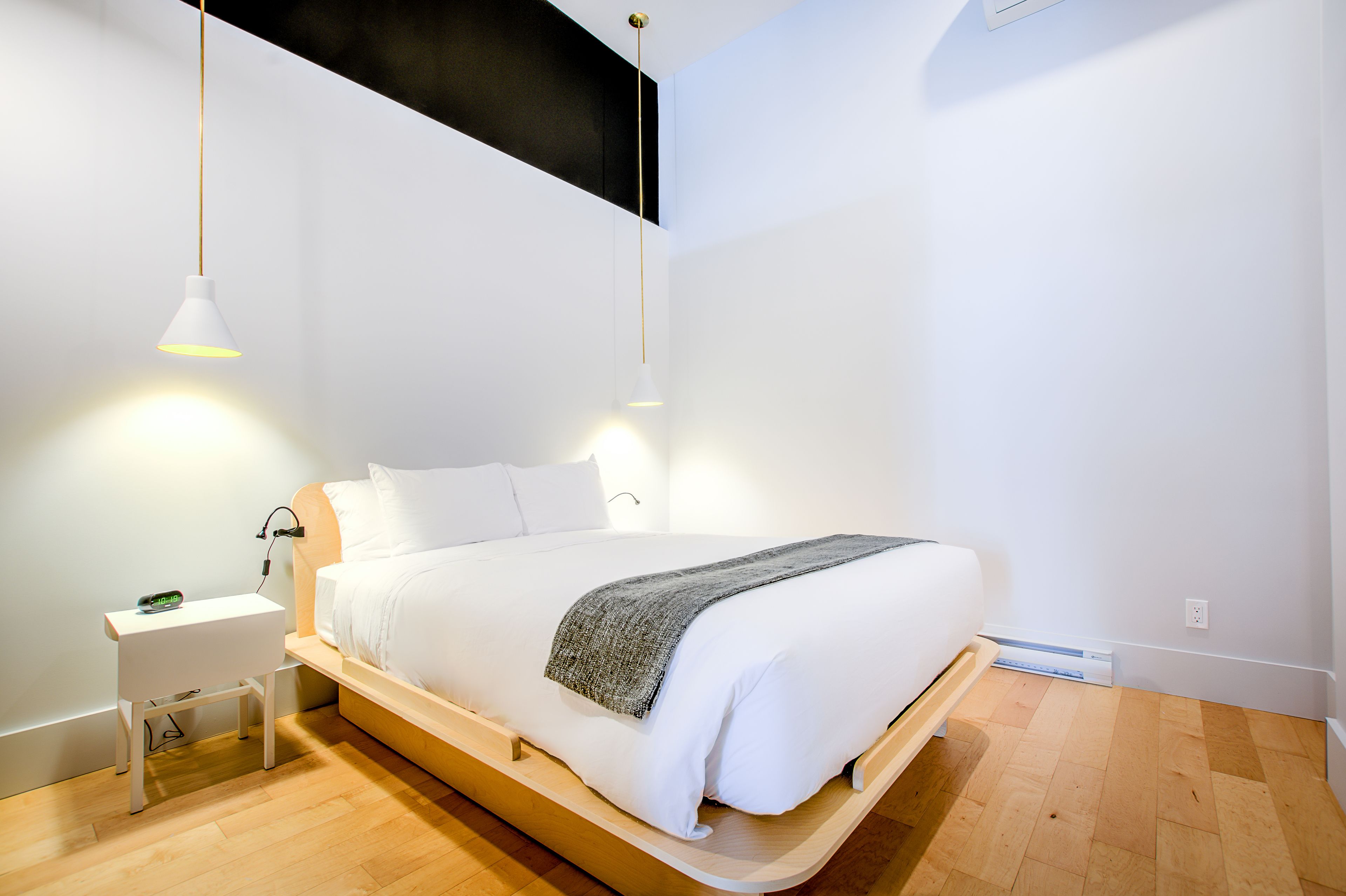 A modern bedroom with a wooden platform bed, white bedding, a bedside table with a lamp, and a power outlet.