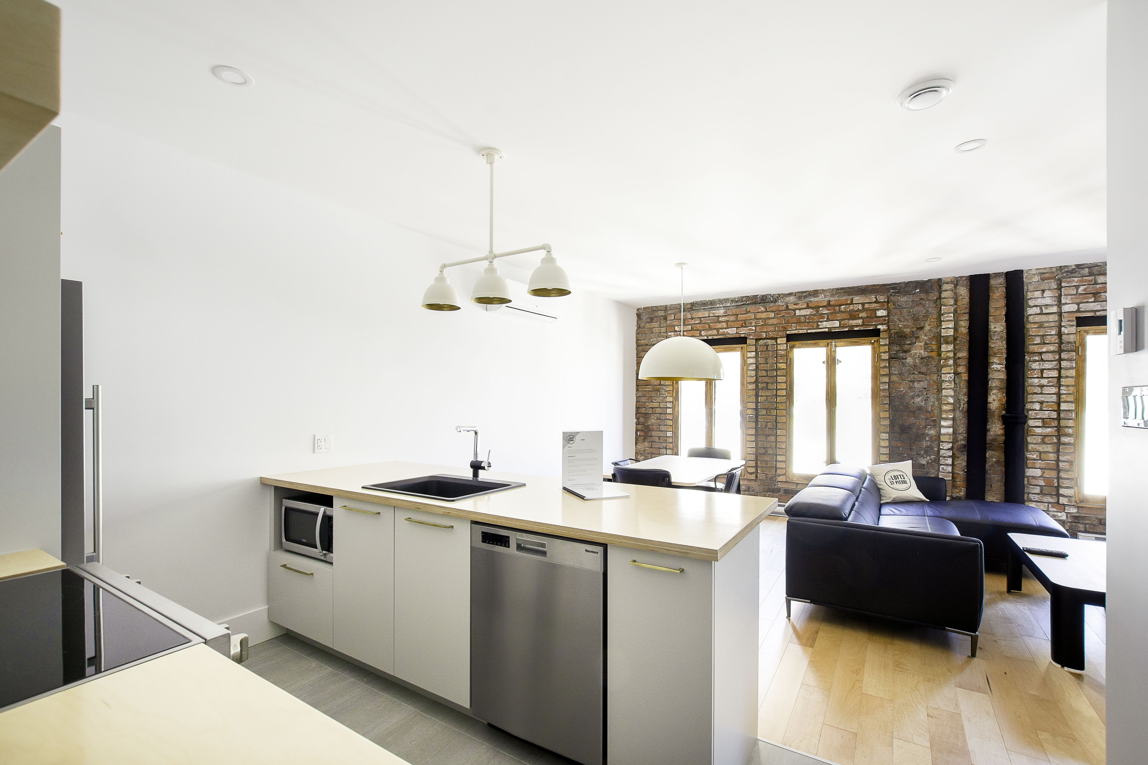A modern kitchen with a built-in dishwasher, a microwave, and a sink. There is a black leather sofa and a white pendant light above the kitchen counter.