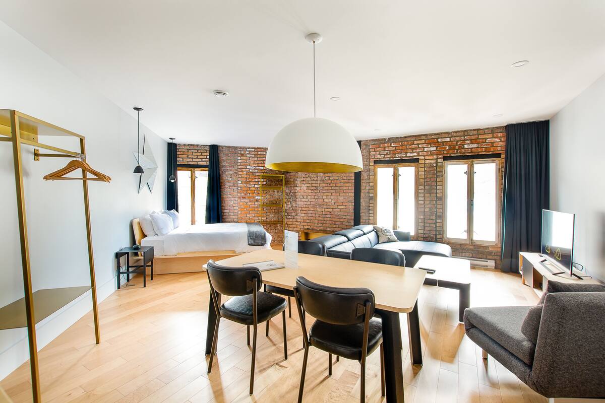 A modern hotel room with a dining area, a sofa, a TV, and a brick wall.