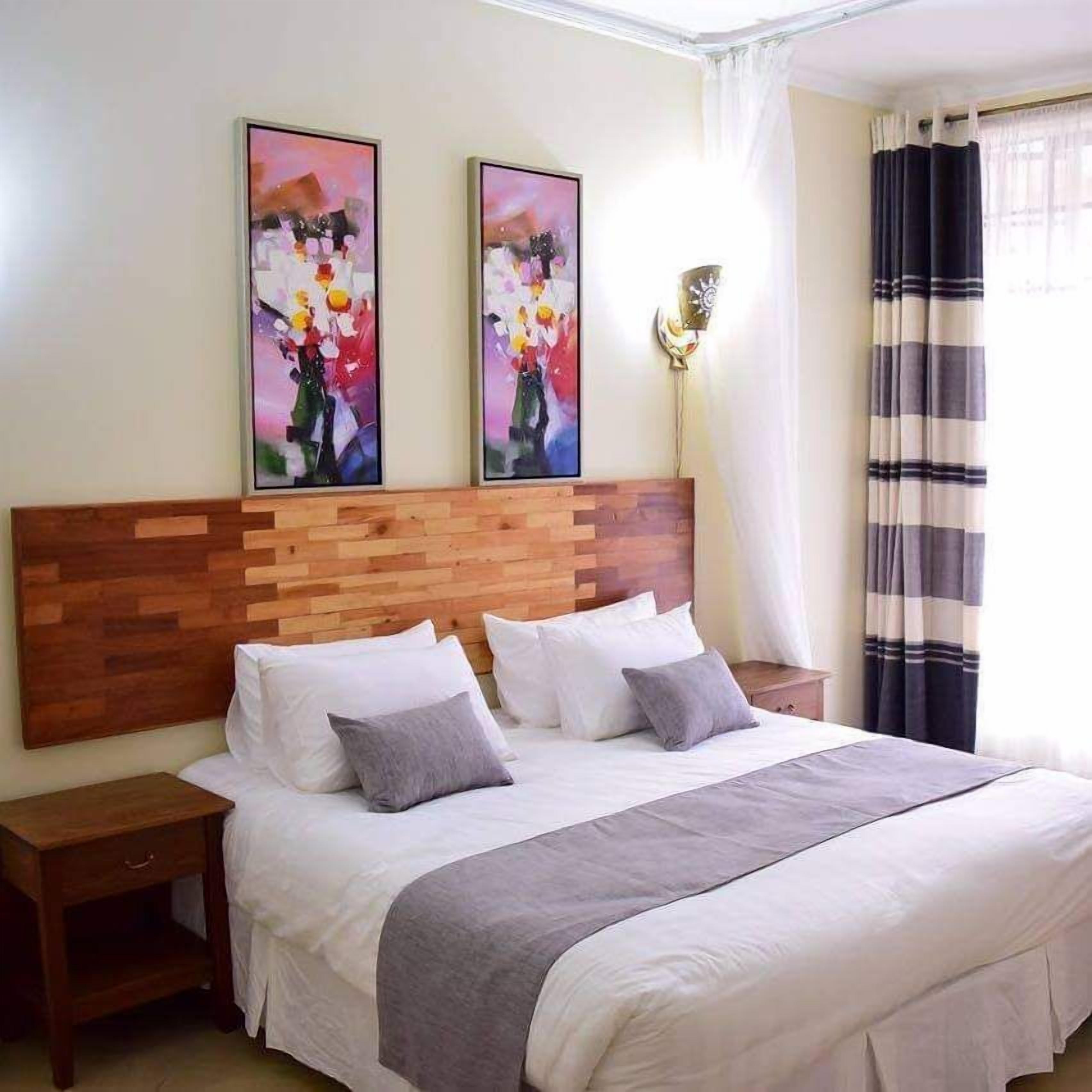 Een hotelkamer met een houten hoofdeinde, een bed met wit en grijs beddengoed, twee schilderijen aan de muur, een nachtkastje en een raam met gordijnen.