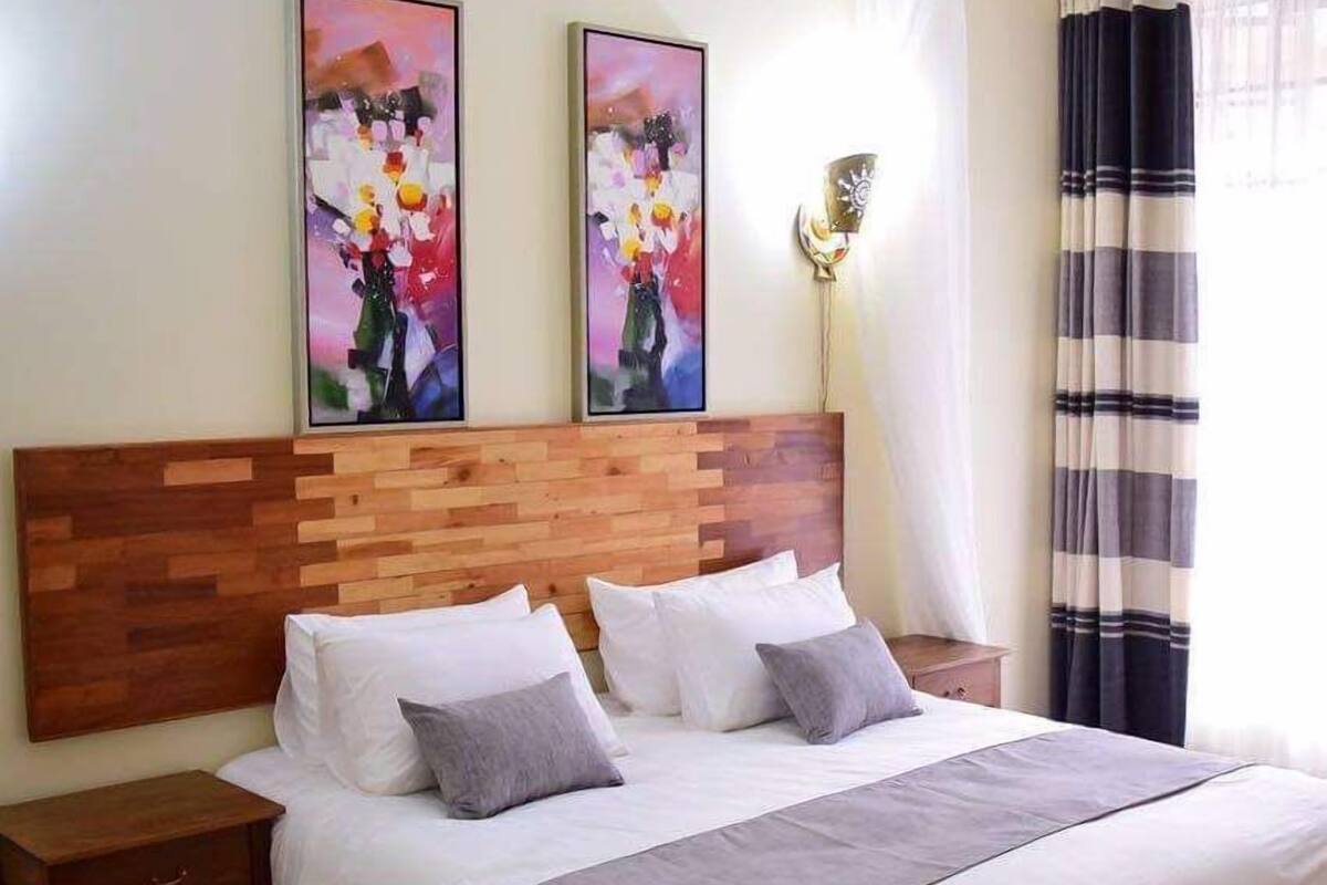 Quarto de hotel com cabeceira de madeira, cama com roupa de cama branca e cinza, duas pinturas na parede, mesa de cabeceira e uma janela com cortinas.