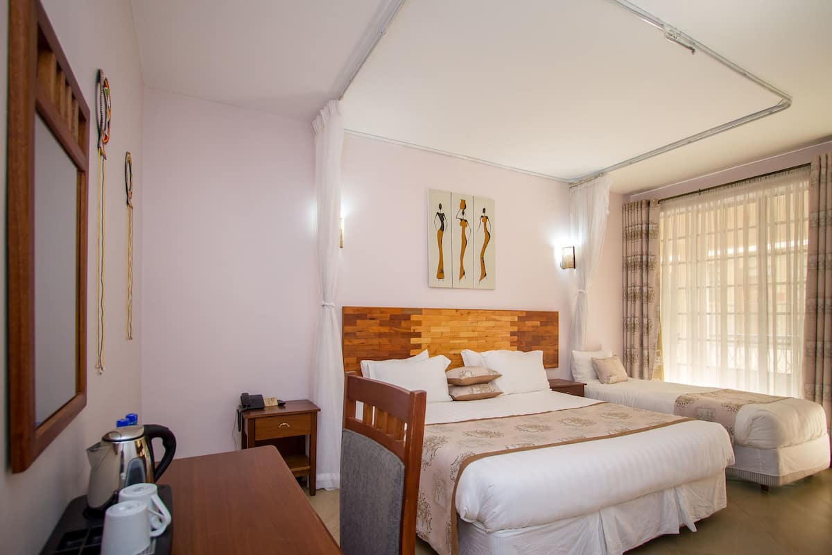 Quarto de hotel com cabeceira de madeira, duas camas, uma escrivaninha de madeira com uma chaleira e uma janela grande com cortinas.